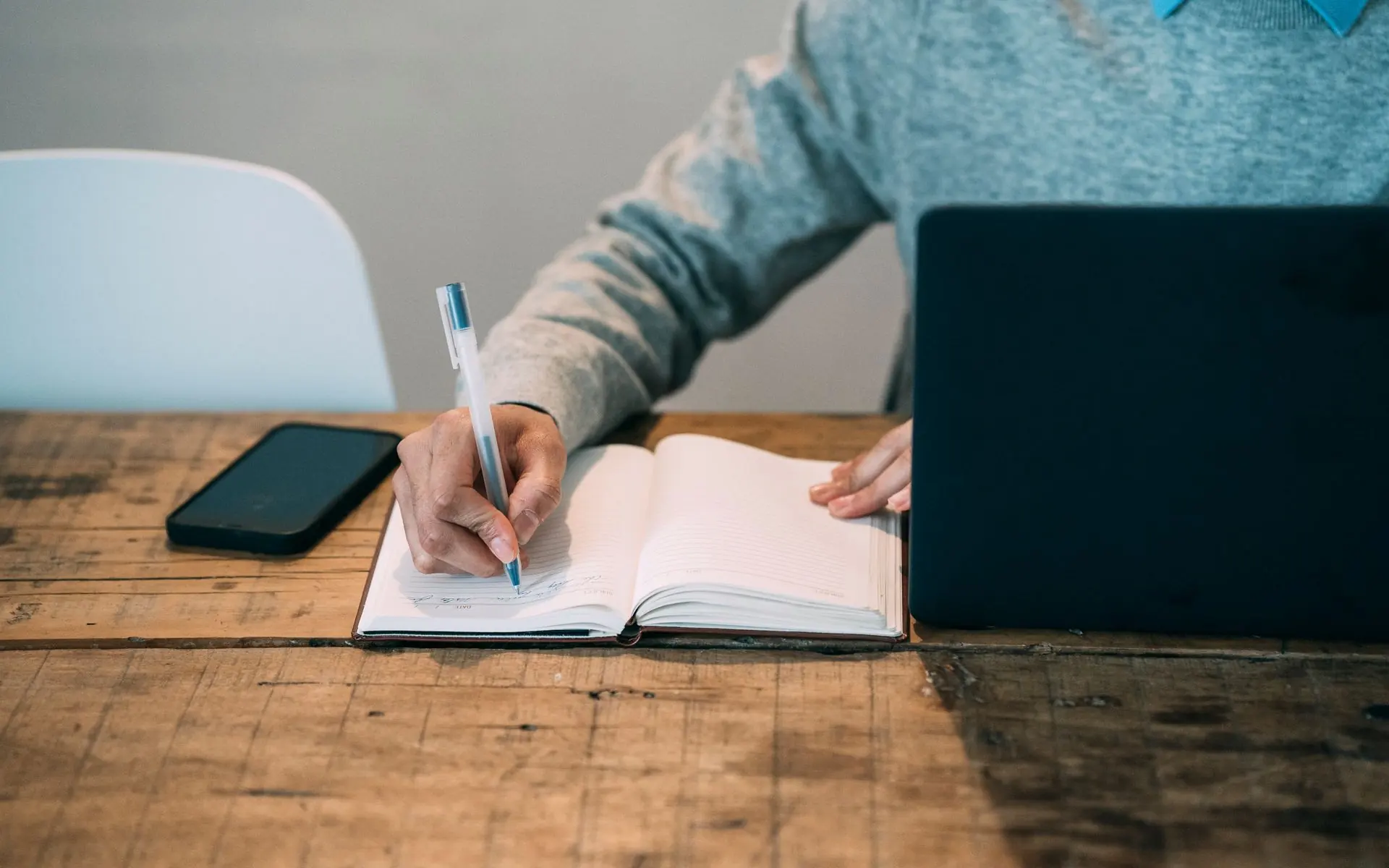 Person writing notes in a planner beside a laptop.