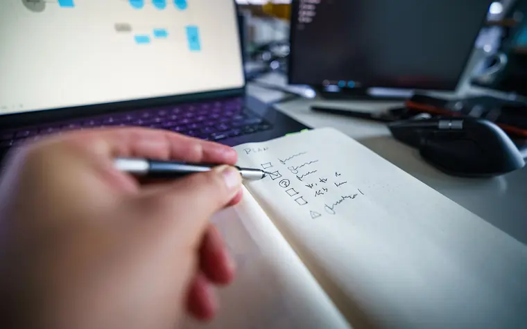 Person writing a checklist in a notebook beside a laptop.