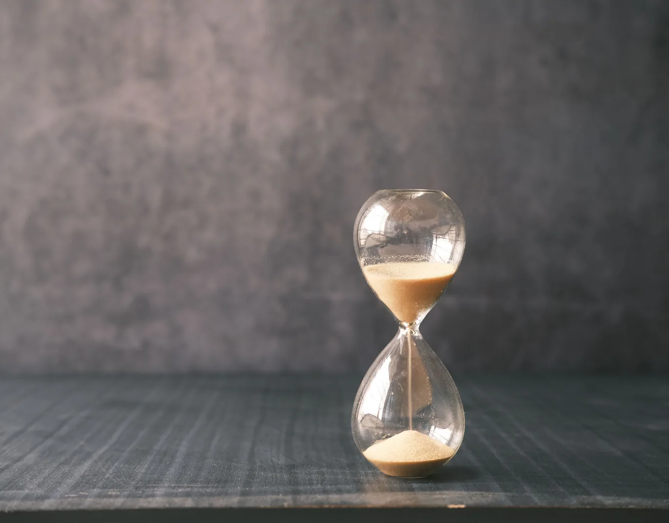 Glass hourglass with sand running through it on a dark timber table against a grey wall.