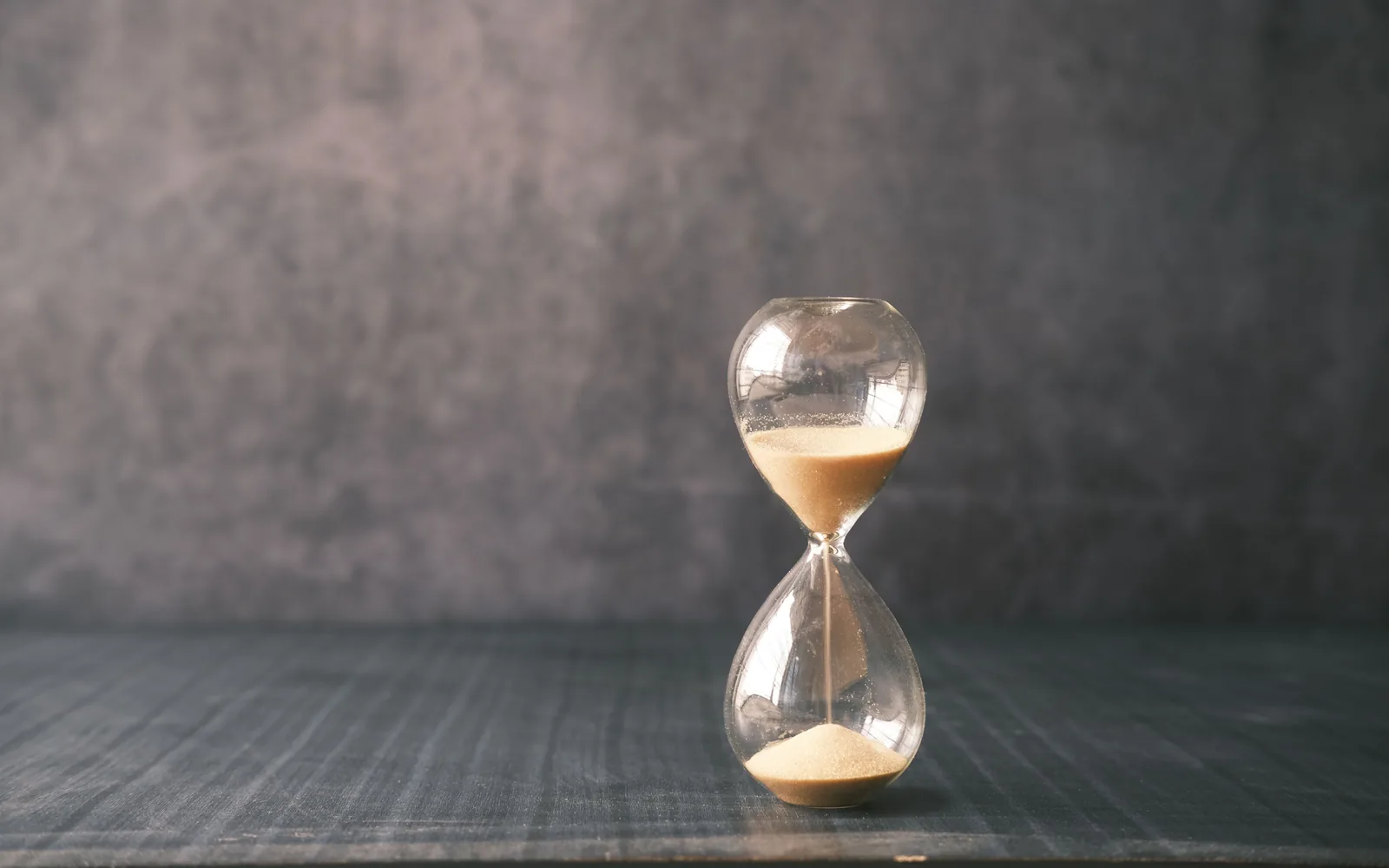 Glass hourglass with sand running through it on a dark timber table against a grey wall.