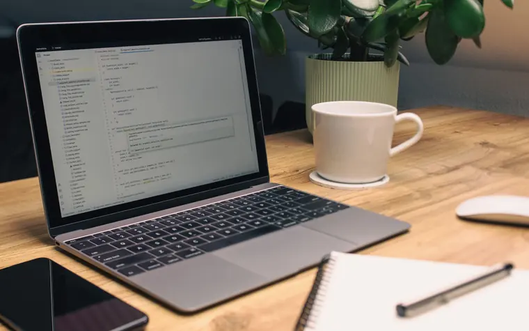 Laptop showing code beside a notebook and coffee.
