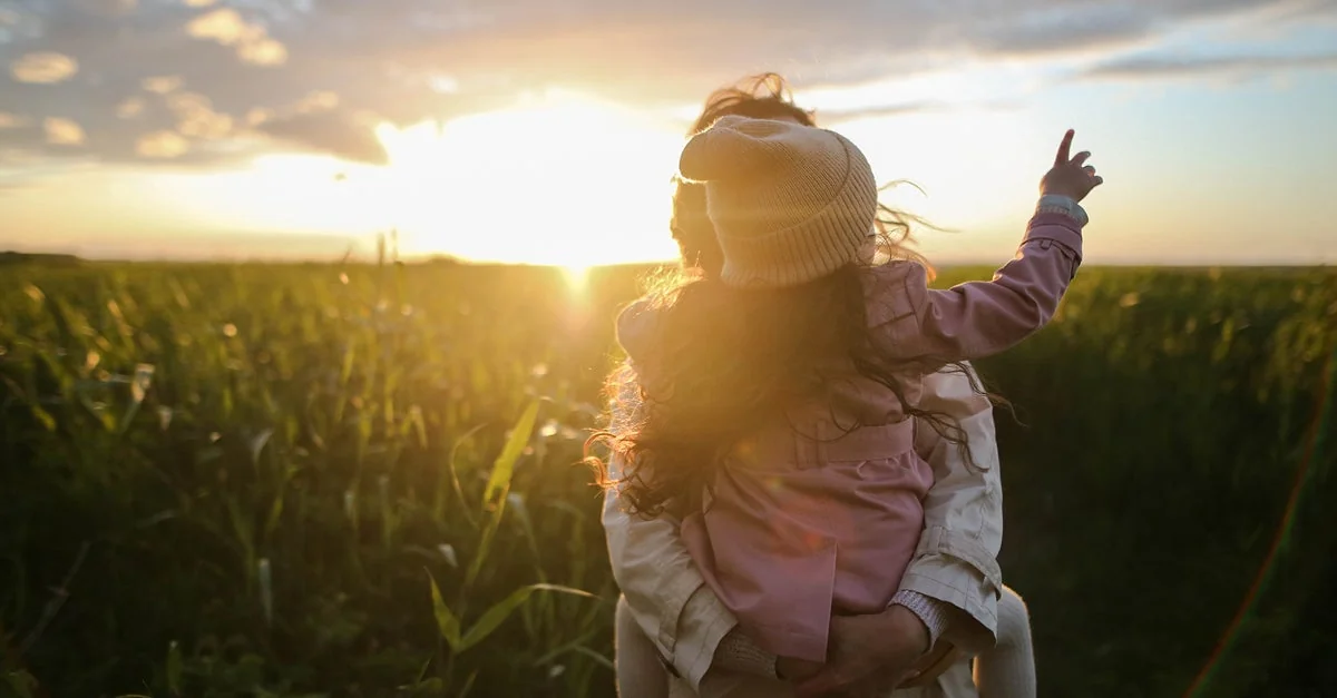 Adult carrying a child through a grassy field.