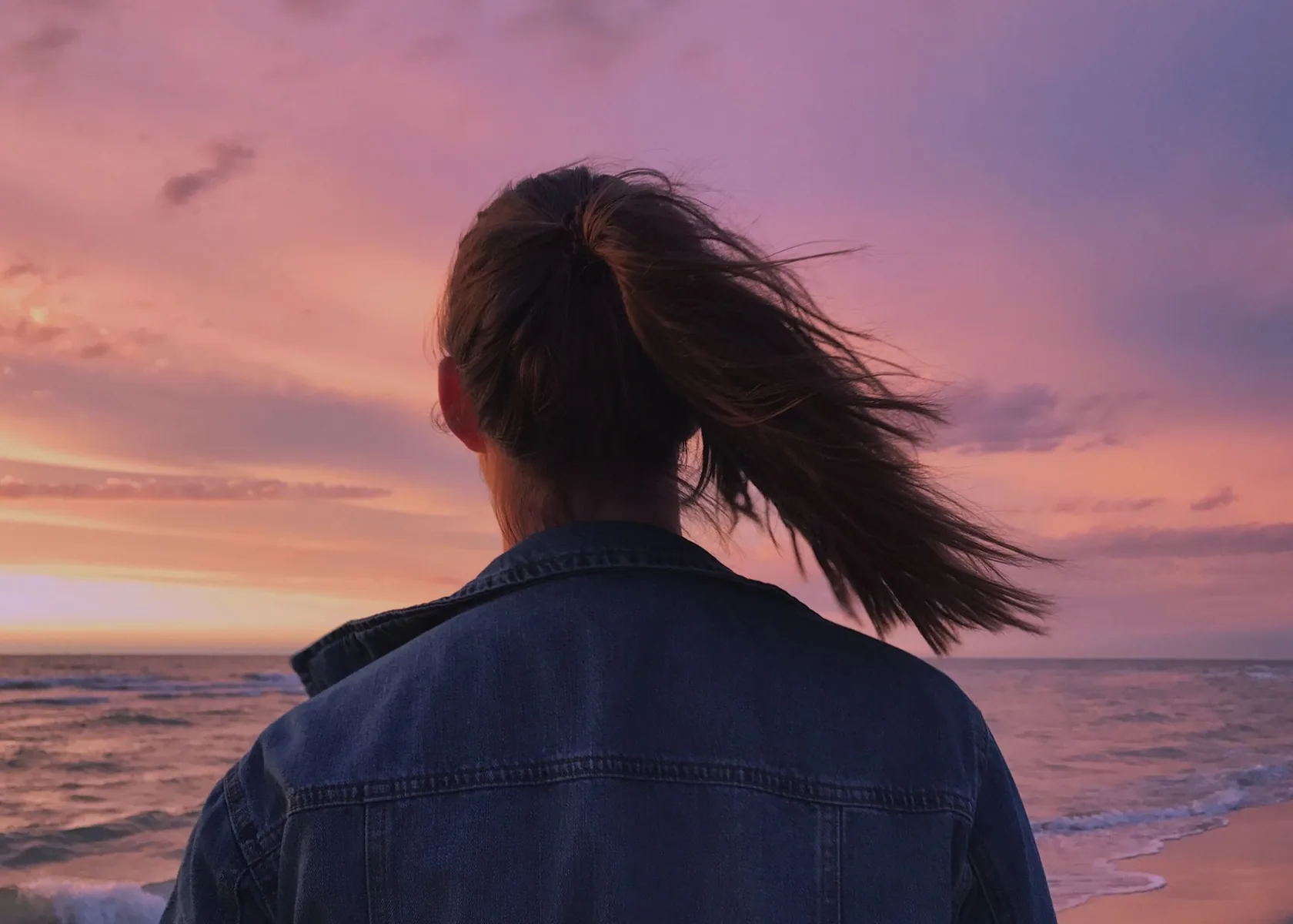 Woman looking out to sea at dusk with her hair lifted by the breeze.
