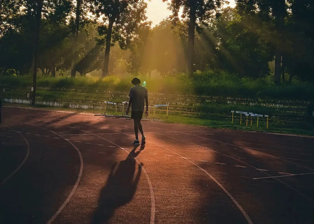 Runner standing on an athletics track at sunrise with light streaming through the trees.